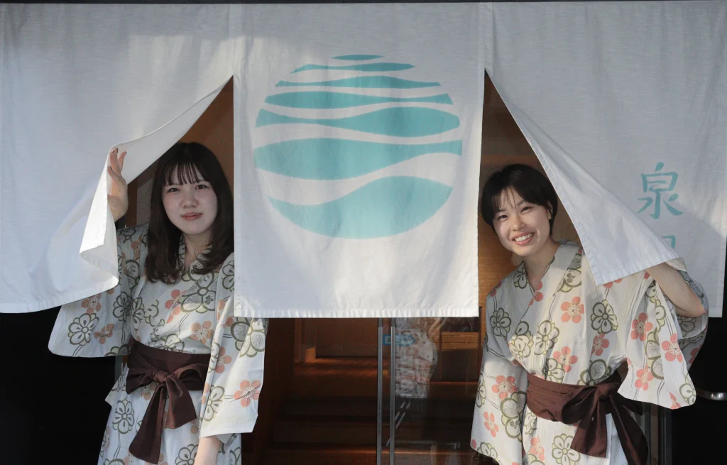 城崎温泉 泉翠の暖簾から、花柄のゆかた姿で顔をのぞかせる女性二人 コピー